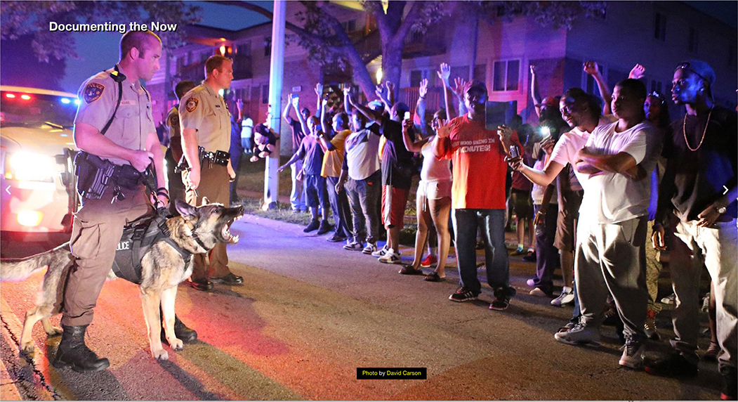 Two police officers stand on a residential street at night, illuminated by the flashing lights of a police vehicle behind them. Across from them, a group of protesters stands in a line on the sidewalk, many with their hands raised.