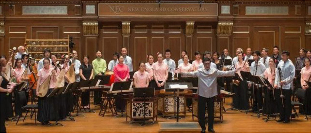 A group of musicians and a conductor on stage at the New England Conservatory, with traditional Chinese instruments visible. The conductor is positioned at the front, gestures with open arms toward the audience.