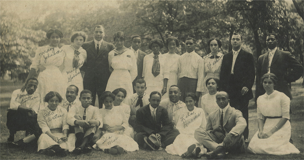 A group of people posing together outdoors. The group consists of approximately 25 persons, arranged in 3 rows. The back row has people standing, the middle row has people seated on the ground, and the front row has individuals sitting cross-legged.