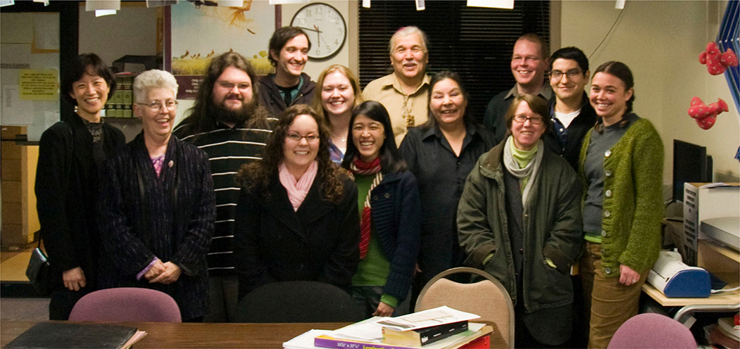 A group of thirteen people smiling and posing together in an indoor office setting. They are dressed in casual and semi-formal attire, with some wearing sweaters, scarves, or jackets.