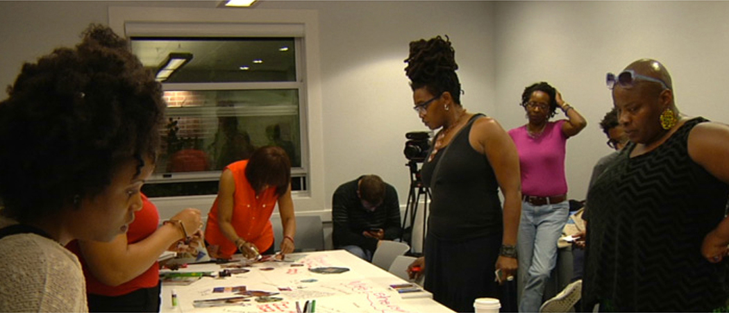 A group of individuals is gathered in a room, standing around a table covered with large sheets of paper, markers, and other creative materials. In the foreground, a woman with curly hair leans over the table, focused on the activity.