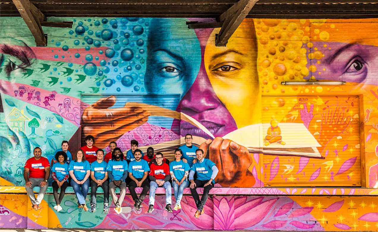 A group of 13 people, wearing matching shirts, sit together on a ledge in front of a large mural. The mural features a close-up of two faces, a hand holding an open book, and various whimsical elements.