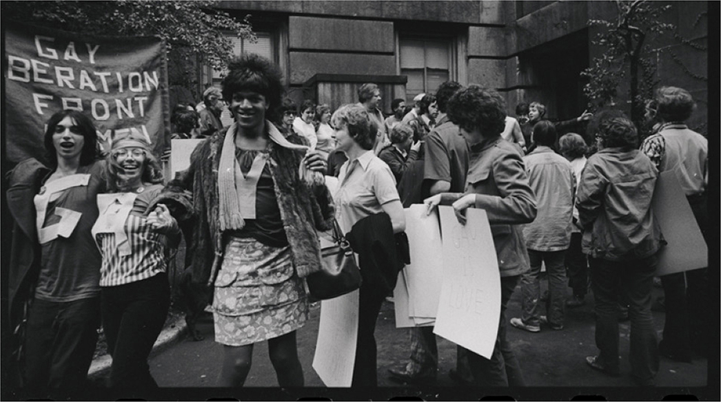 A group of people at a protest, with a banner reading “Gay Liberation Front” in the background. In the foreground, three individuals stand arm-in-arm, smiling and engaging with the camera.
