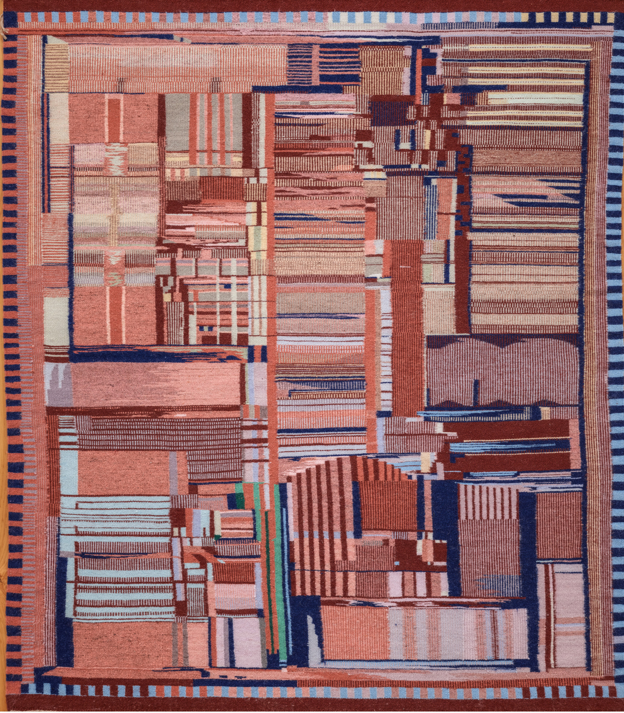 An abstract woven textile featuring a geometric pattern of intersecting lines, rectangles, and grids in varying shades of red, pink, blue, and beige, bordered by alternating striped designs.