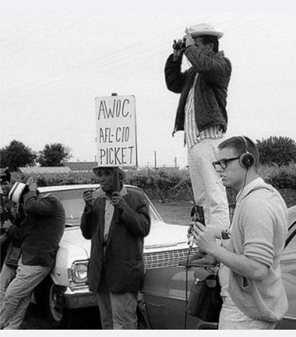 A group of people gathered near a car. A man in the center holds a sign that reads, “A W O C, A F L-C I O PICKET.” He is speaking into a microphone. On the right, a man stands on the car’s hood, using binoculars.