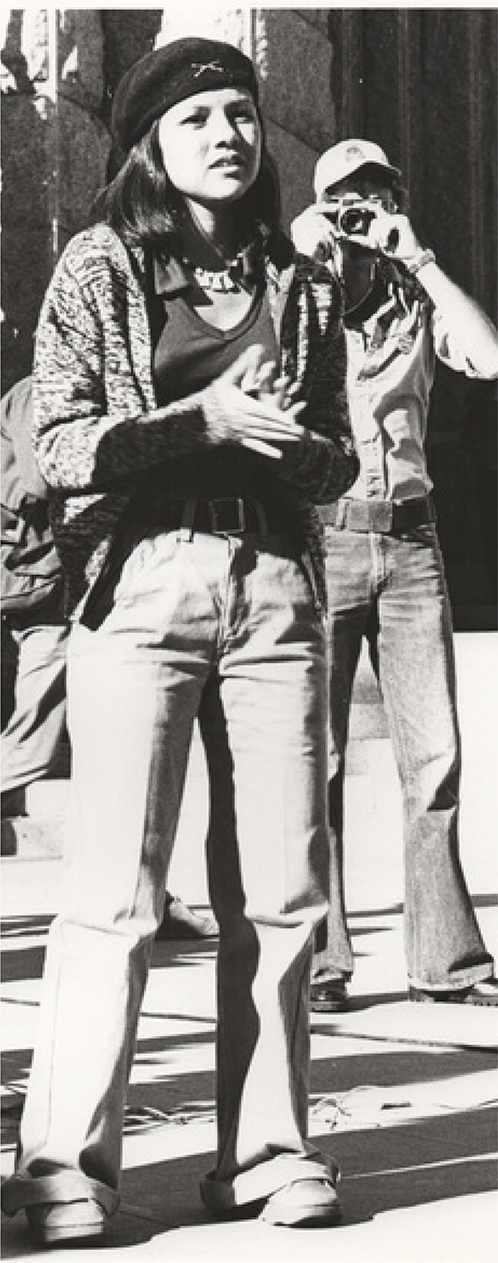 A young woman standing outdoors, speaking or gesturing with her hands. Behind her, a person wearing a cap and sunglasses holds a camera.